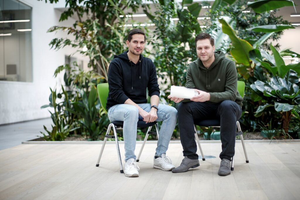 Aufnahme der Gründer von „Green Elephant Biotech“: Felix Wollenhaupt (links) und Dr. Joel Eichmann (rechts). Sie sitzen vor einer grünen Pflanzenkulisse in einem Gebäude.