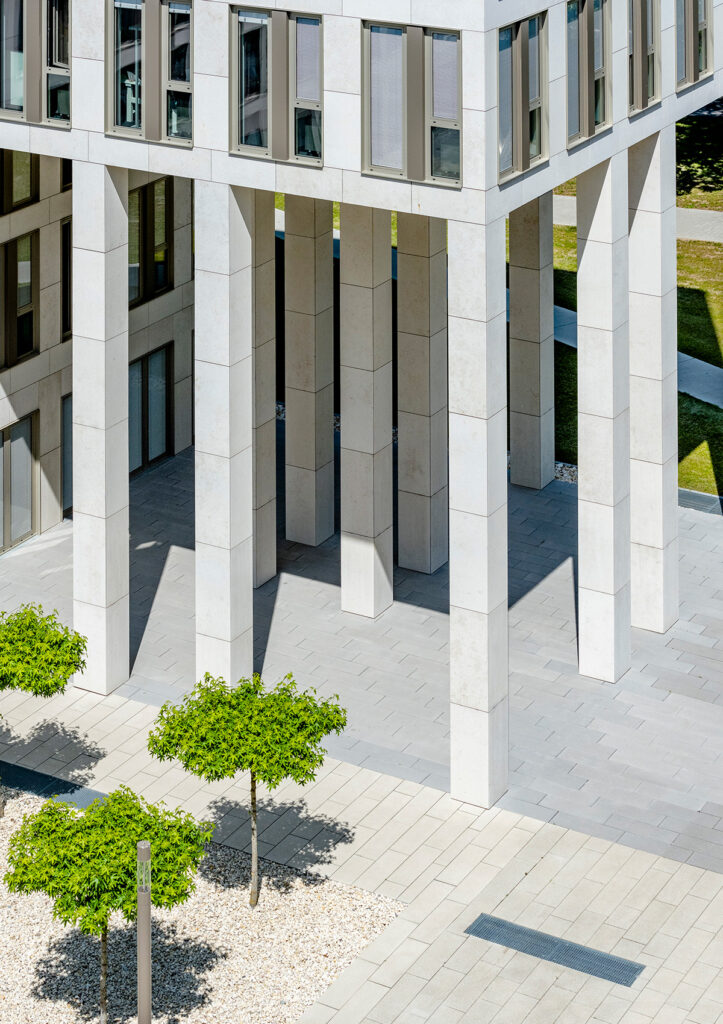Blick von oben auf das BMH-Bürogebäude, das auf mehreren hohen, hellen Säulen ruht, davor gepflasterter Platz mit jungen Bäumen und Kiesfläche.
