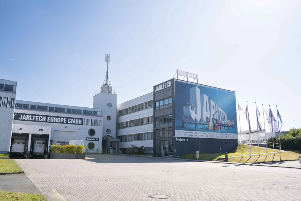 Das mehrstöckige Firmengelände von „Jarltech“: Eingangsbereich und Fassade mit großem Banner mit „Jarltech“-Schriftzug, dazu Fahnenmasten und Lagerhalle mit Verladetoren.