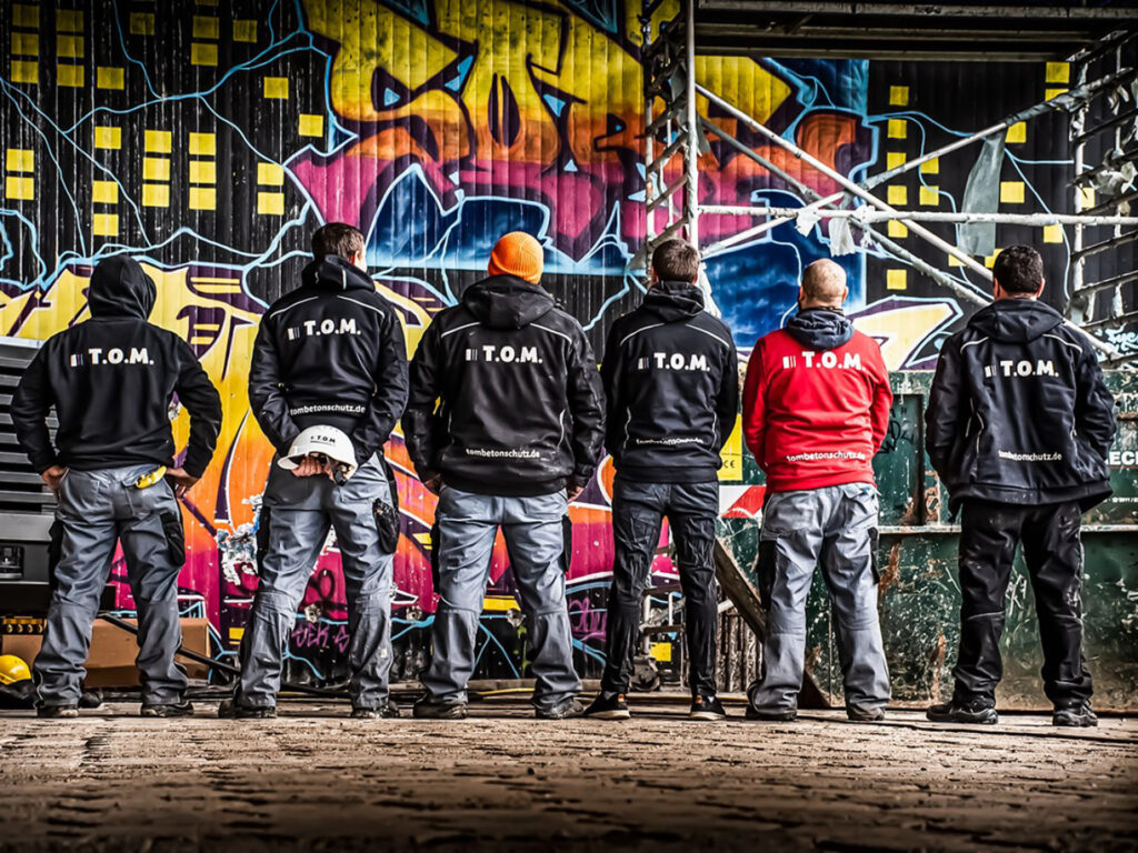 Gruppenaufnahme des Teams von „T.O.M.“: Sechs Männer stehen mit dem Rücken zur Kamera vor einer bunten Graffitiwand, daneben ein Baugerüst und ein Container.