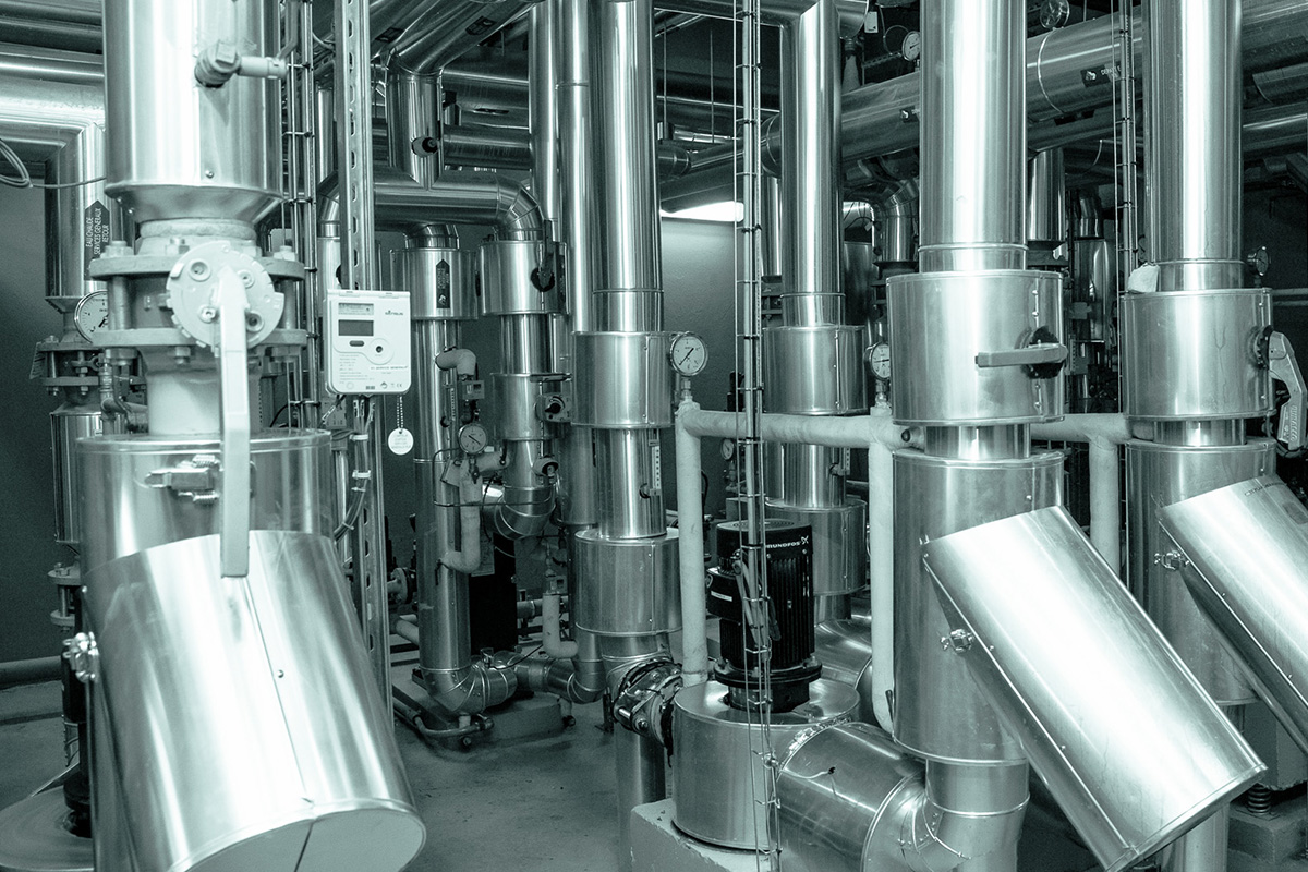 Industrielle Anlage mit zahlreichen Edelstahlrohren, Tanks, Ventilen, Pumpen und Messinstrumenten in einem technischen Betriebsraum.