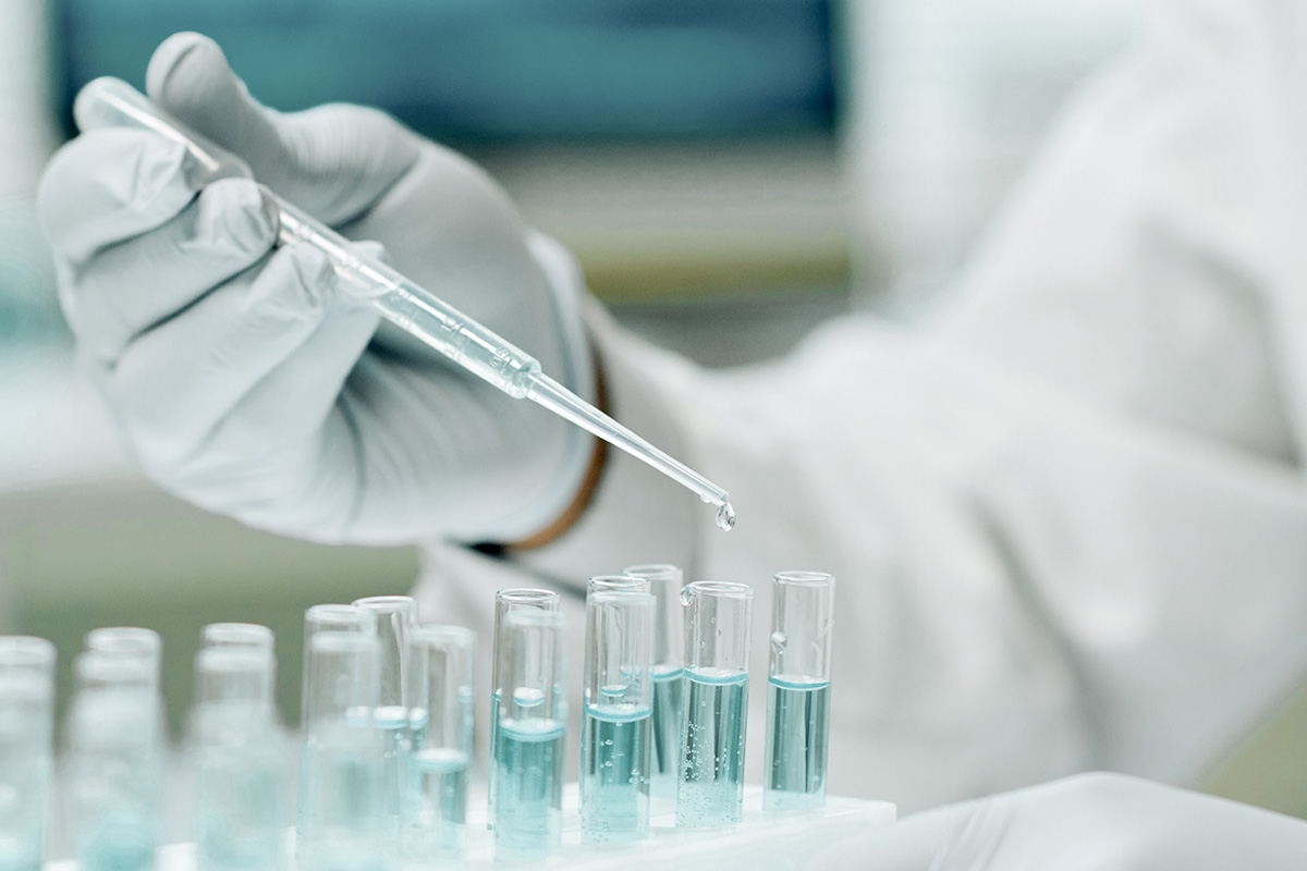 Eine Handschuhtragende Person hält eine Pipette über mehrere Reagenzgläser mit blauer Flüssigkeit in einem Labor.
